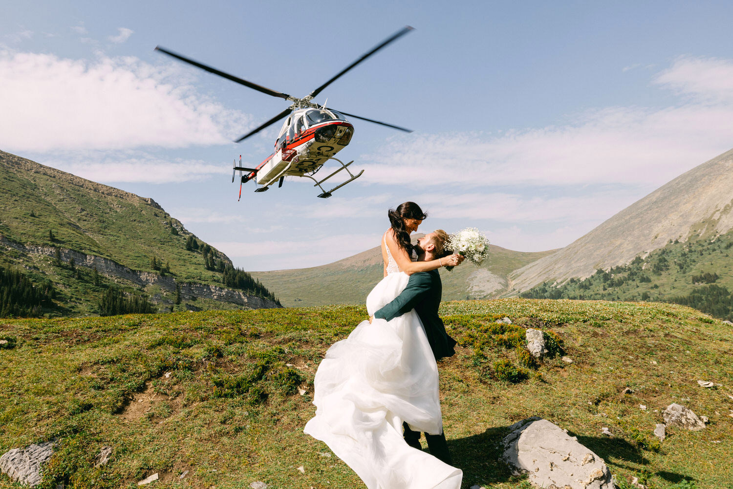 A couple celebrates their wedding outdoors, with a helicopter flying overhead against a backdrop of mountains and lush greenery.