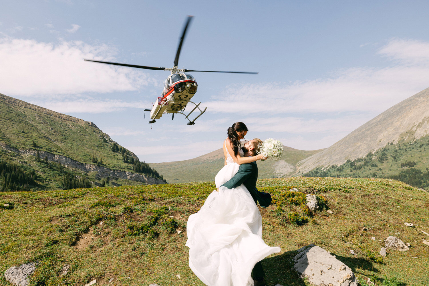 A joyful couple embraces on a mountainside with a helicopter flying overhead.