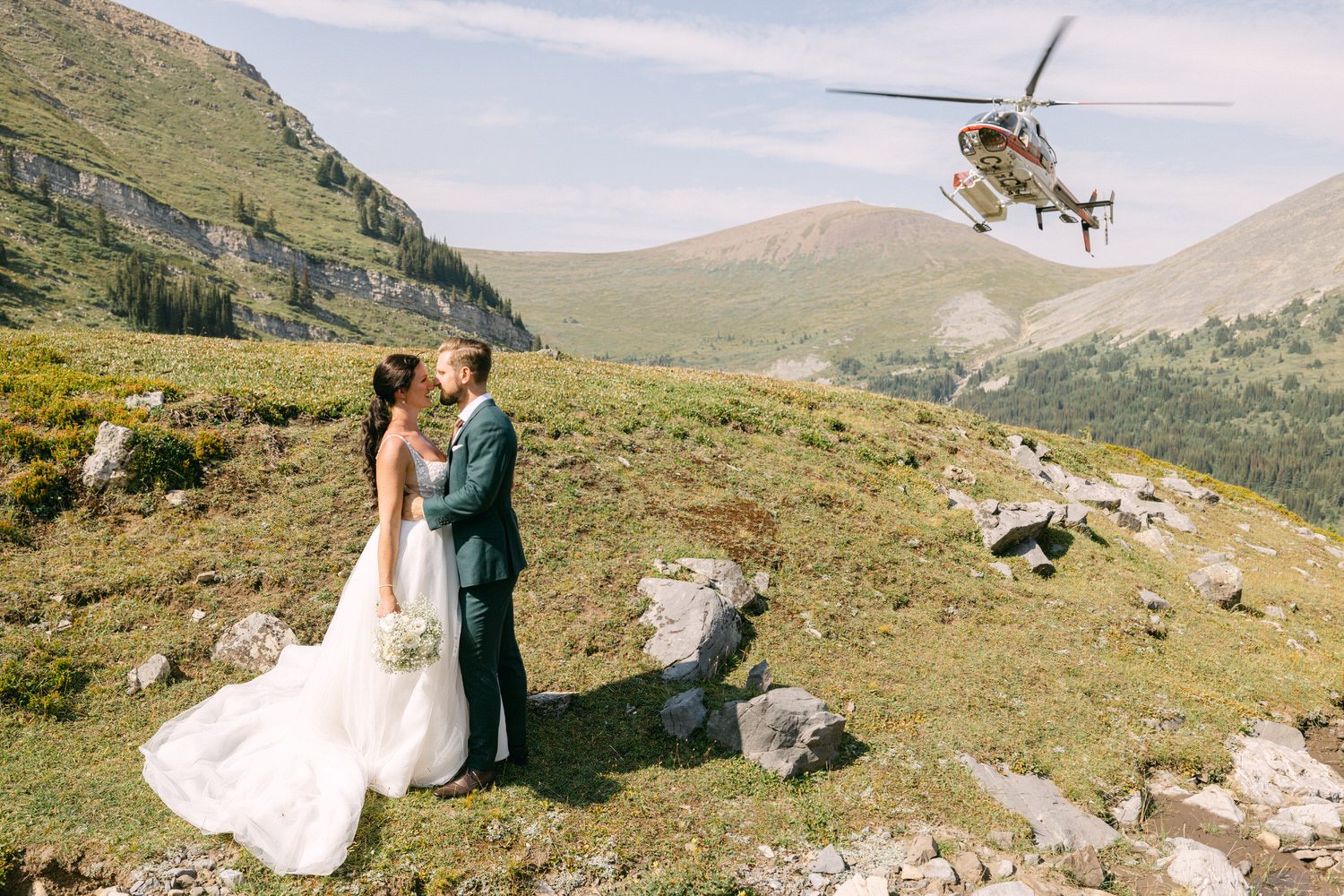 A bride and groom share a romantic moment amidst stunning mountain scenery, with a helicopter flying overhead.