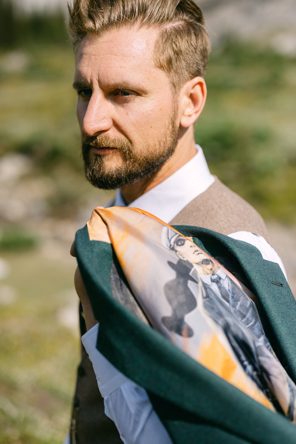 A close-up of a well-groomed man holding a jacket over his shoulder, revealing a colorful inner lining, against a scenic outdoor backdrop.