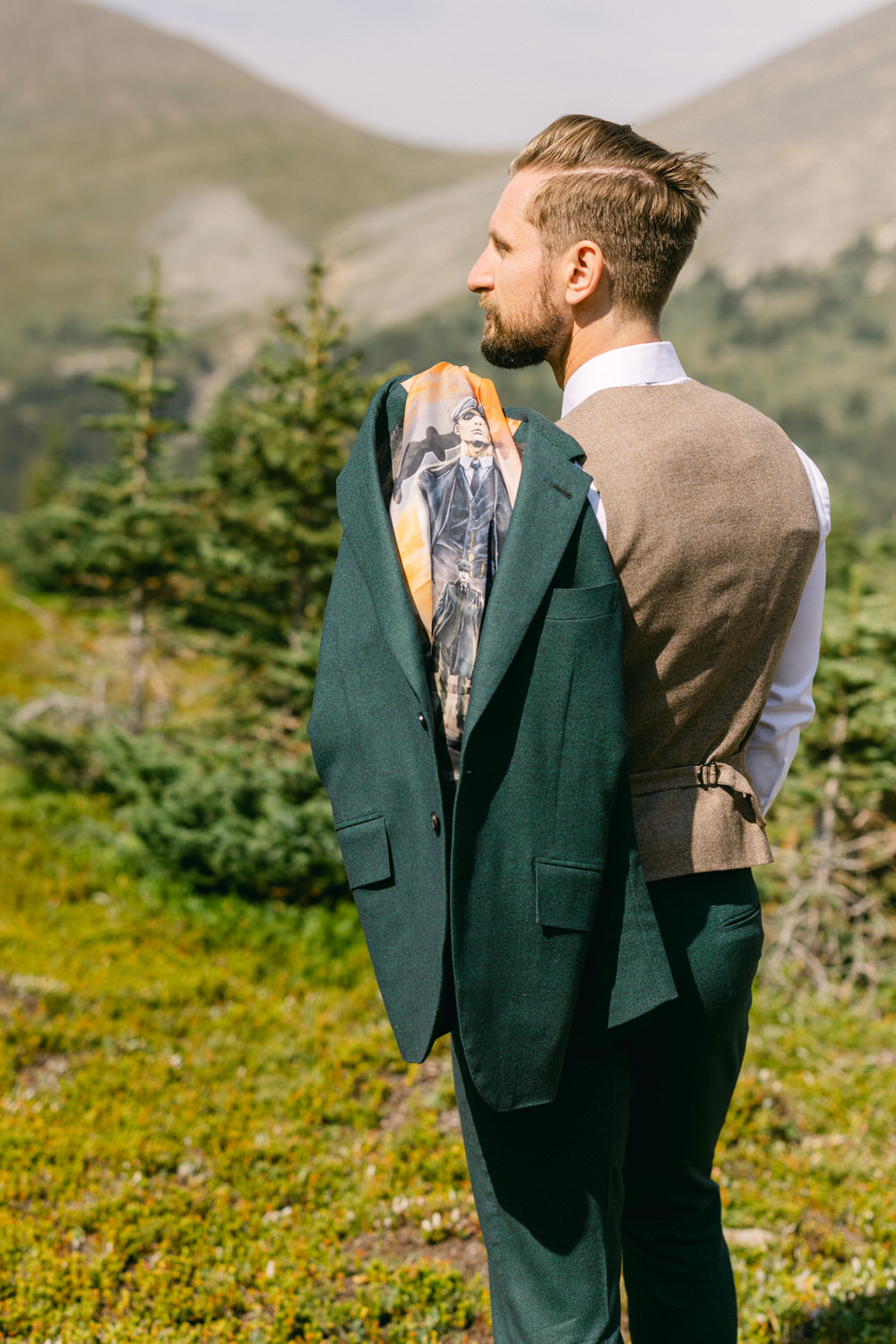 A man dressed in a green suit jacket and tan vest stands outdoors, holding a jacket with a colorful design on its lining, against a backdrop of mountains and trees.