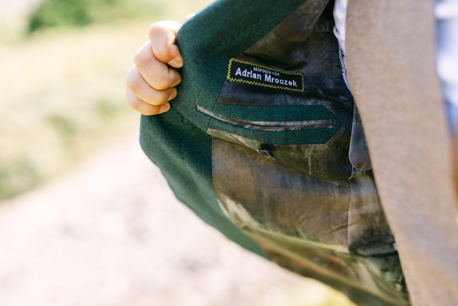 A close-up of a bespoke suit's inner label, showcasing the name "Adrian Mroczek" with a green and gray fabric background.