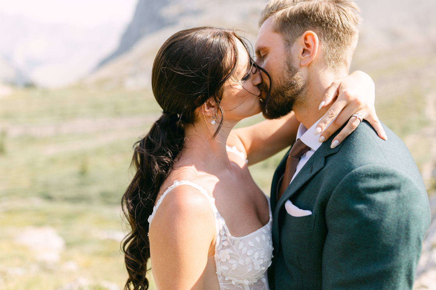 A couple shares a tender kiss in a scenic outdoor setting, surrounded by nature and mountains.