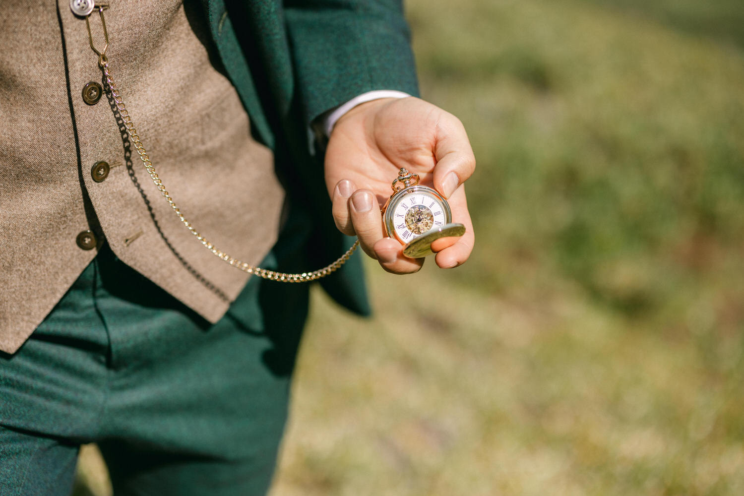 A close-up of a person holding a vintage pocket watch with a decorative chain, dressed in a stylish green suit and brown vest, set against a blurred natural background.