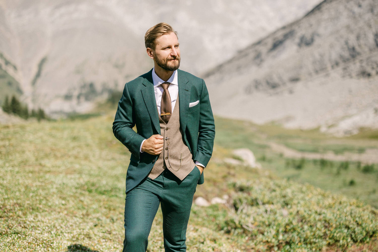 A well-dressed man in a green suit with a vest walks confidently through a mountainous landscape, showcasing a blend of elegance and nature.