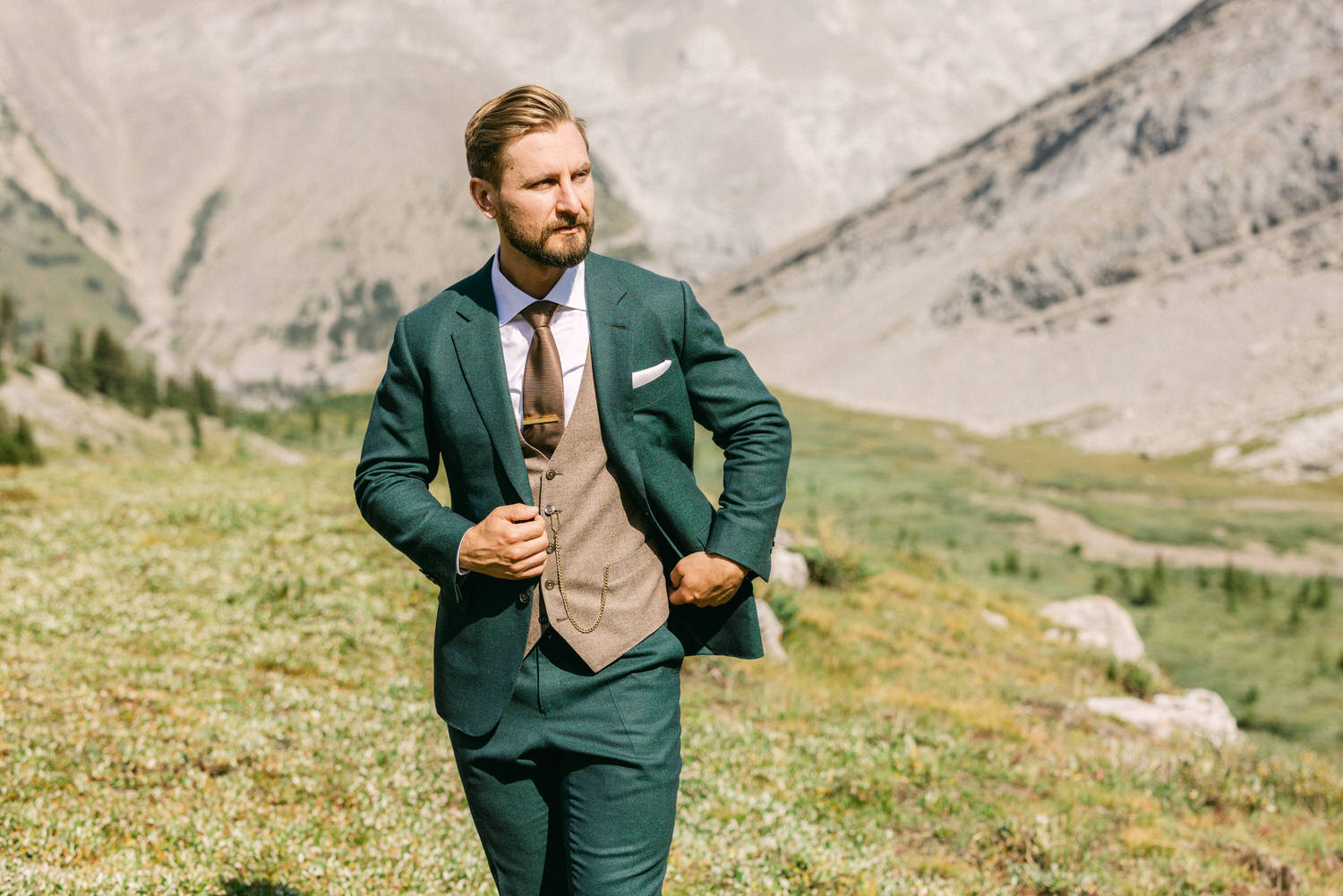 A well-dressed man in a green suit and vest stands confidently in a mountainous landscape, showcasing a blend of elegance and nature.