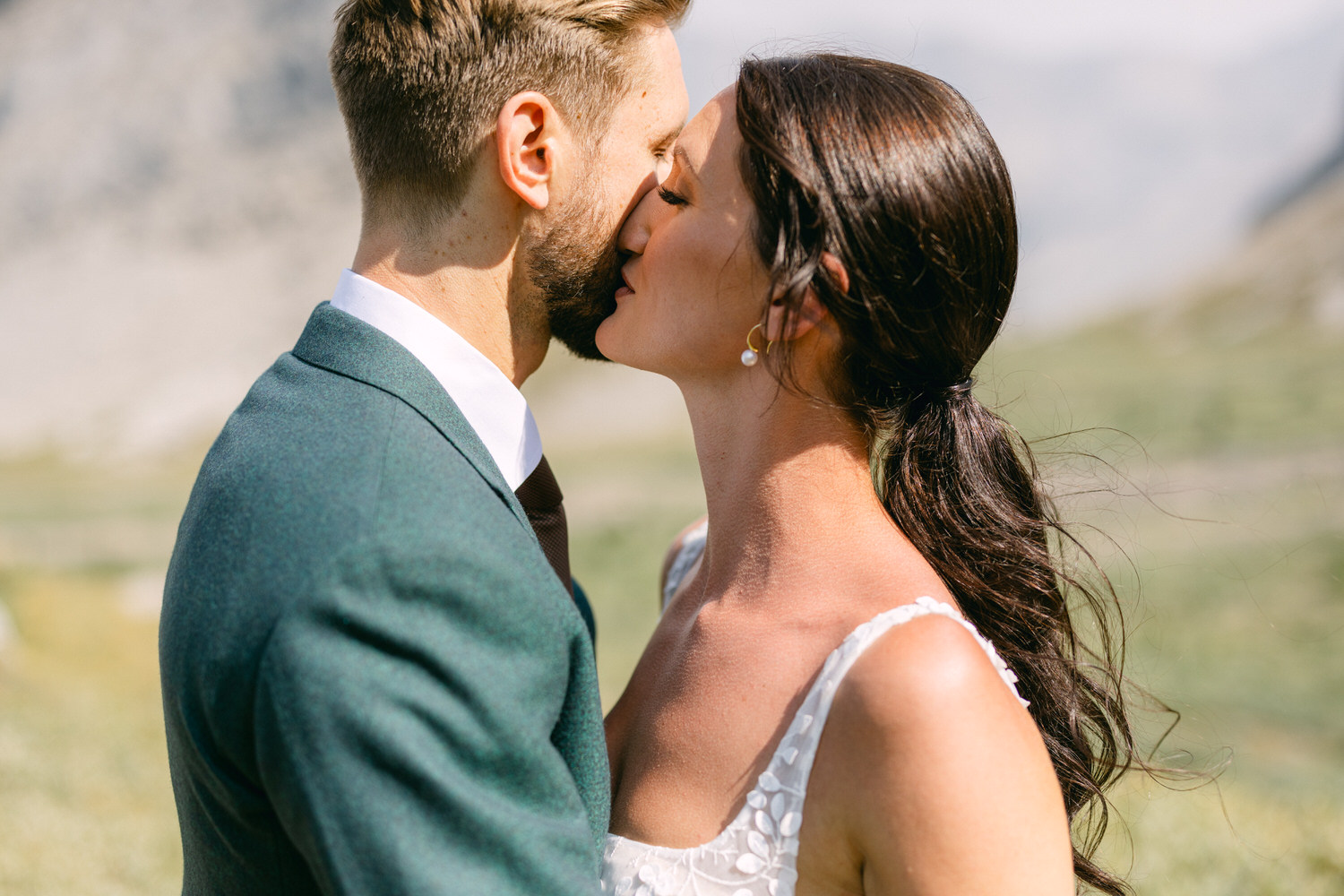 A couple sharing a tender kiss in a scenic outdoor setting, featuring a lush green background and the groom in a green suit while the bride wears a delicate dress, capturing a moment of love and intimacy.