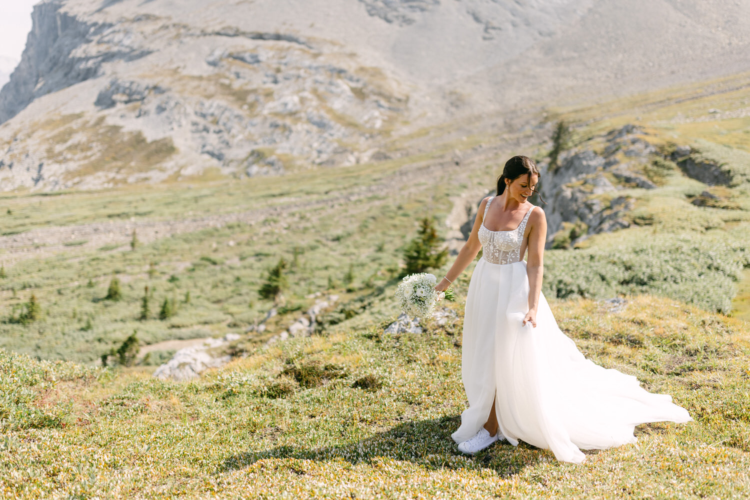 A bride wearing a flowing white dress and sneakers, holding a bouquet, stands on a lush green hillside surrounded by mountains.