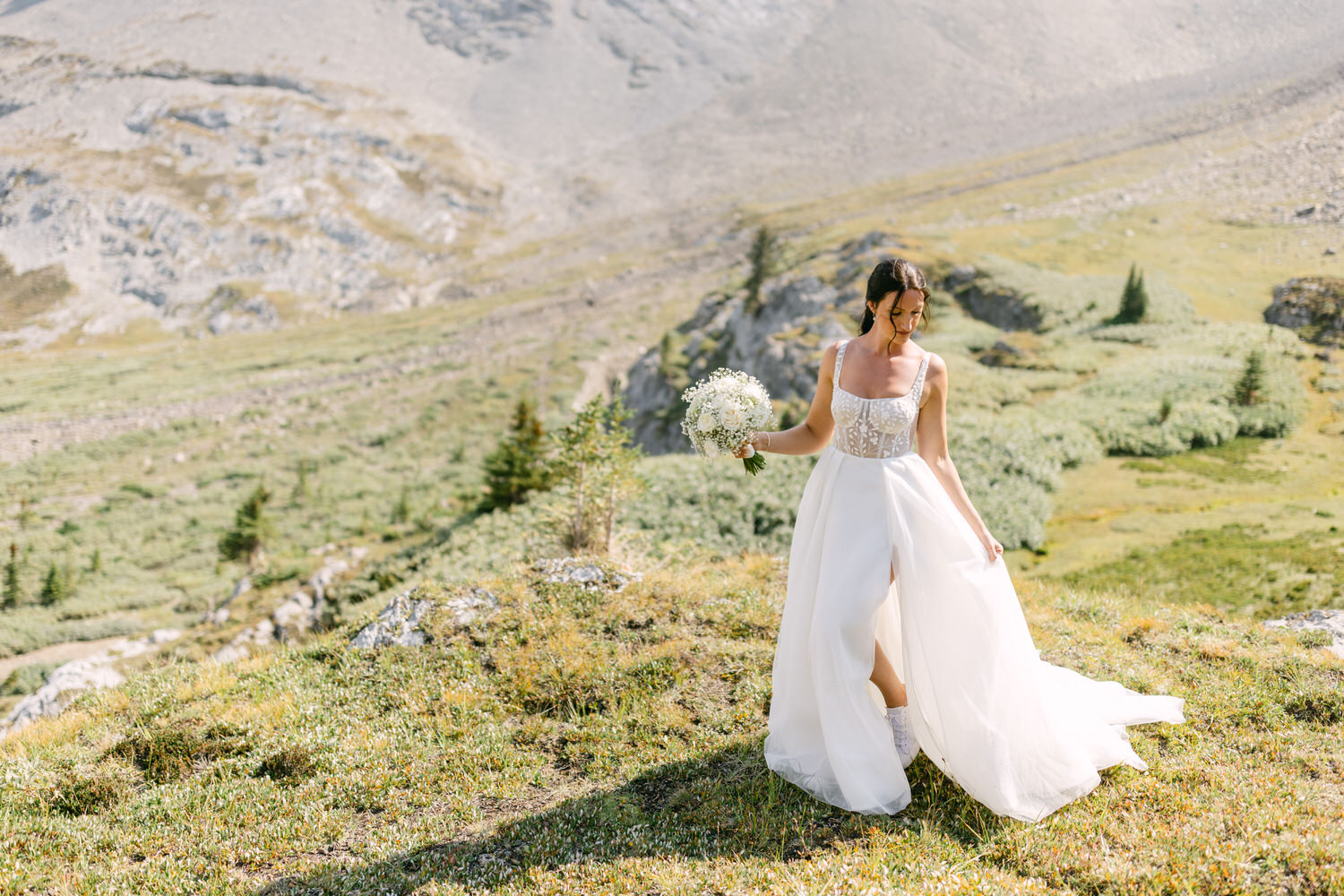 A bride in a flowing white dress holds a bouquet while walking through a lush mountain landscape with greenery and rocky terrain in the background.