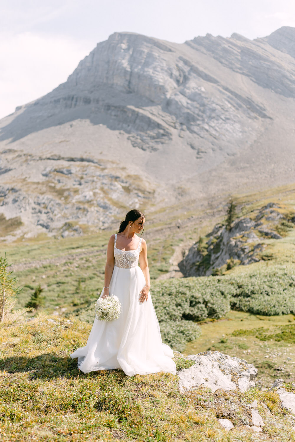 A bride in a flowing white gown holding a bouquet stands on a hillside with stunning mountain scenery in the background.