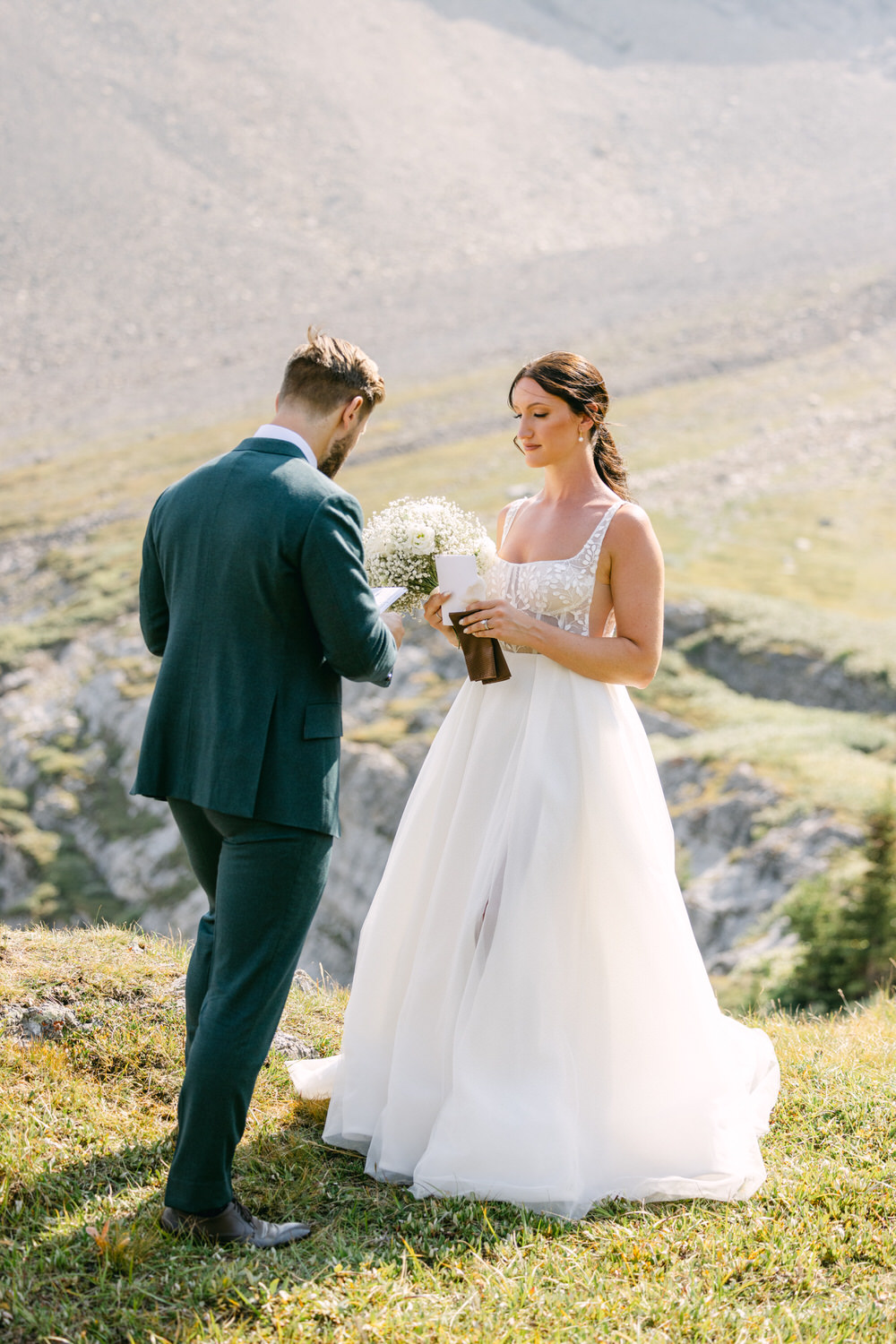 A bride and groom exchange vows in a scenic outdoor setting, surrounded by mountains and greenery. The bride holds a bouquet while the groom reads from a note.