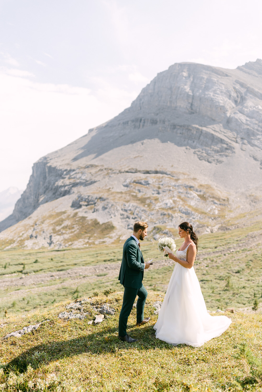 A bride in a flowing white gown holds a bouquet while exchanging vows with a groom in a dark suit against a stunning mountainous backdrop.