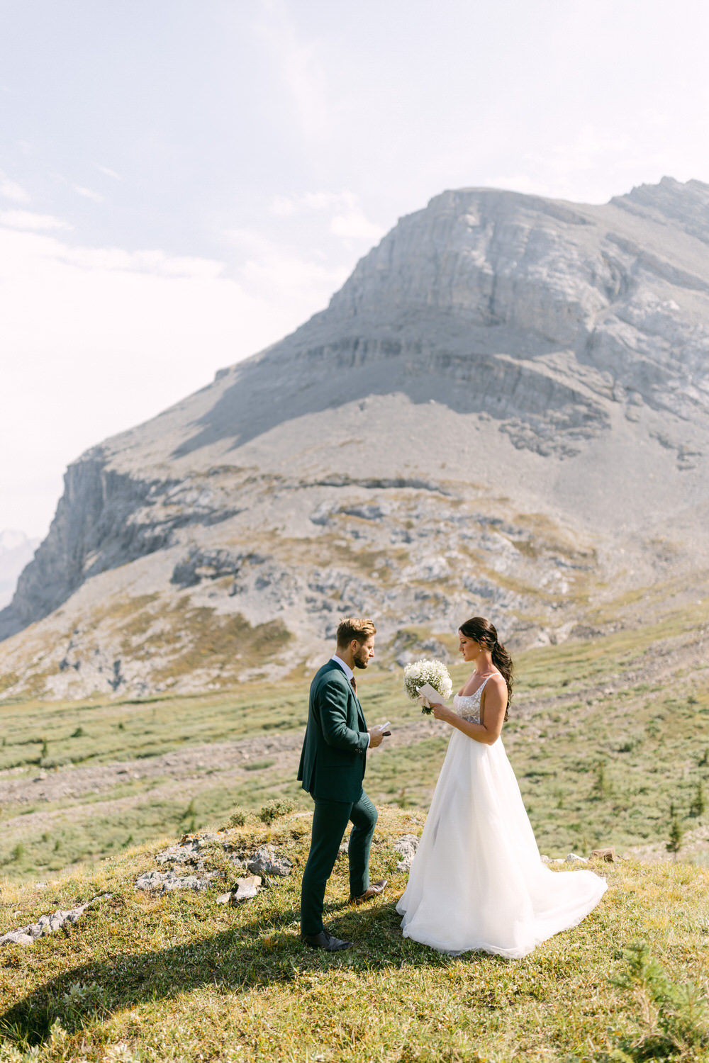 A bride in a flowing white gown holds a bouquet while exchanging vows with a groom in a green suit amidst a stunning mountainous backdrop.