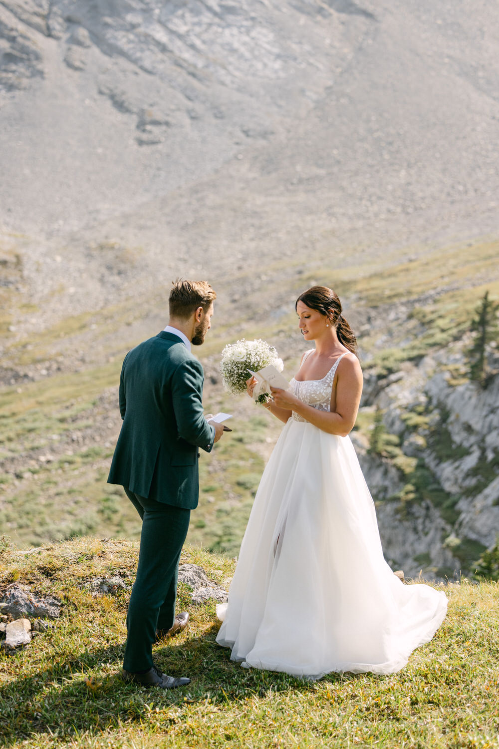 Mountain Wedding Ceremony::A couple exchanges vows on a grassy hilltop, surrounded by stunning mountain scenery, with the bride holding a bouquet and the groom reading from a card.