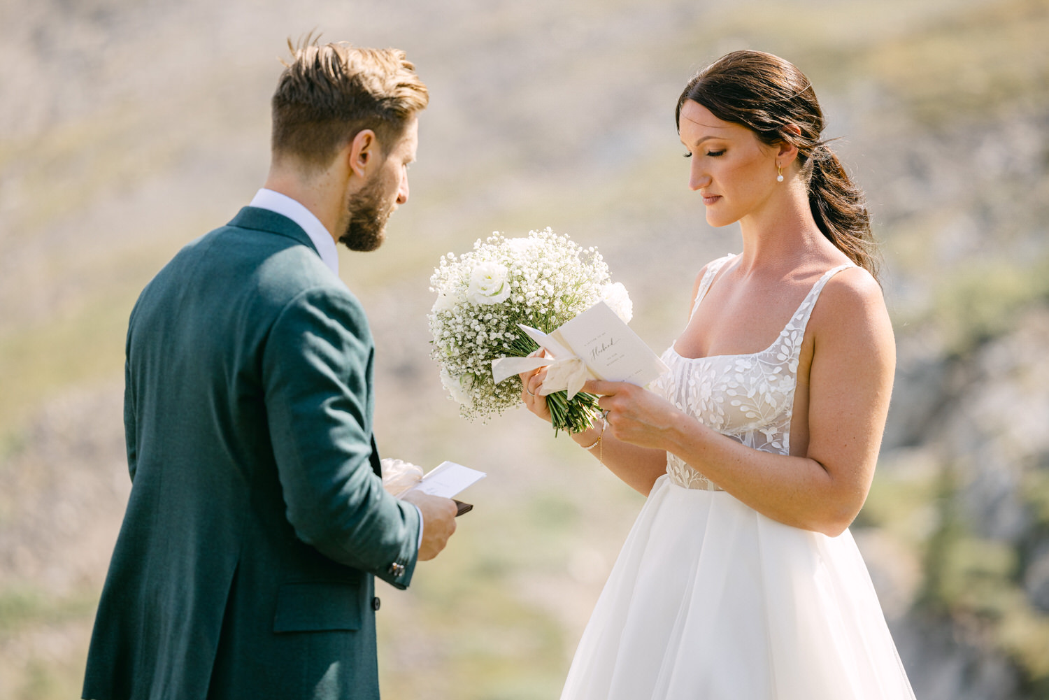 A bride reads her vows while holding a bouquet, as her partner listens attentively during an outdoor wedding ceremony.