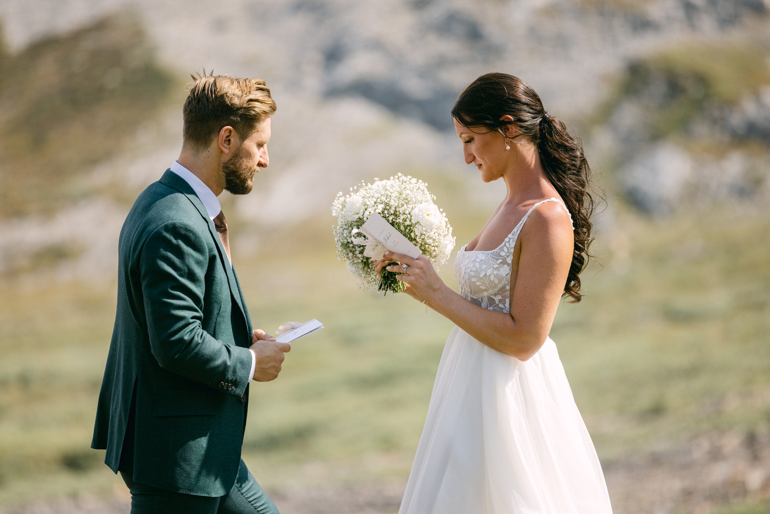 A bride and groom exchanging vows in a picturesque outdoor setting, with the bride holding a bouquet and reading from a card.