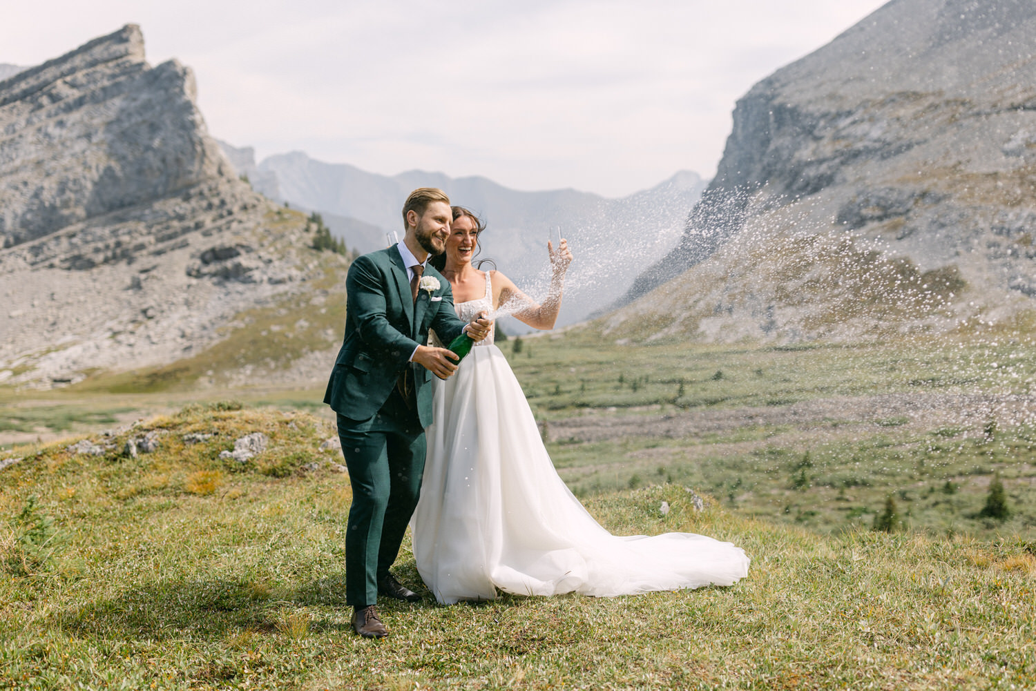 A bride and groom joyfully popping champagne in a scenic mountainous landscape, surrounded by lush greenery and dramatic peaks.