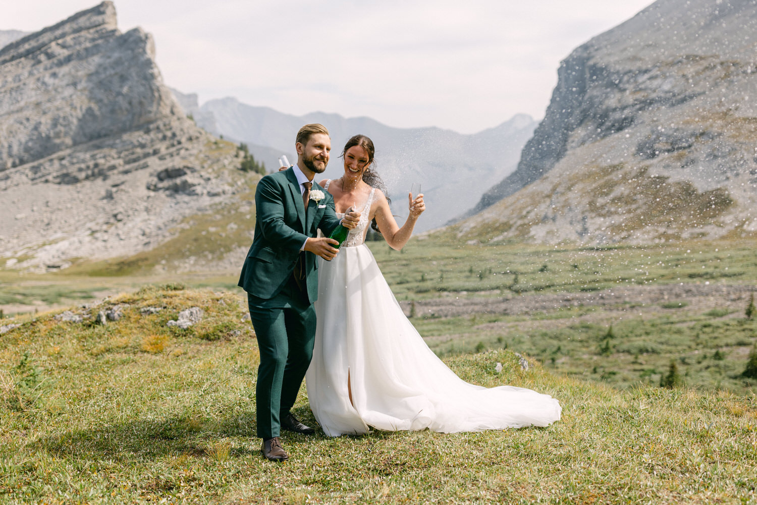 A newlywed couple joyfully popping a champagne bottle on a grassy hillside, surrounded by mountains, with sparkling wine splashing around them.