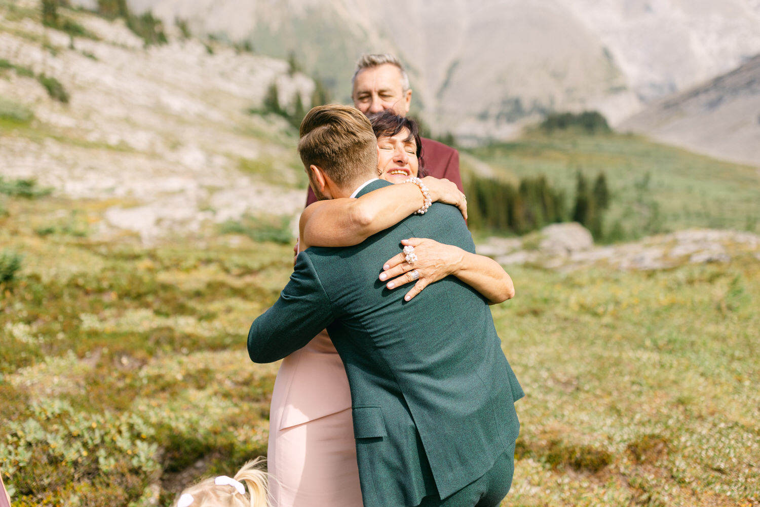 A heartfelt moment capturing a man embracing a woman in a scenic outdoor setting, with a joyful expression on her face and another person in the background.