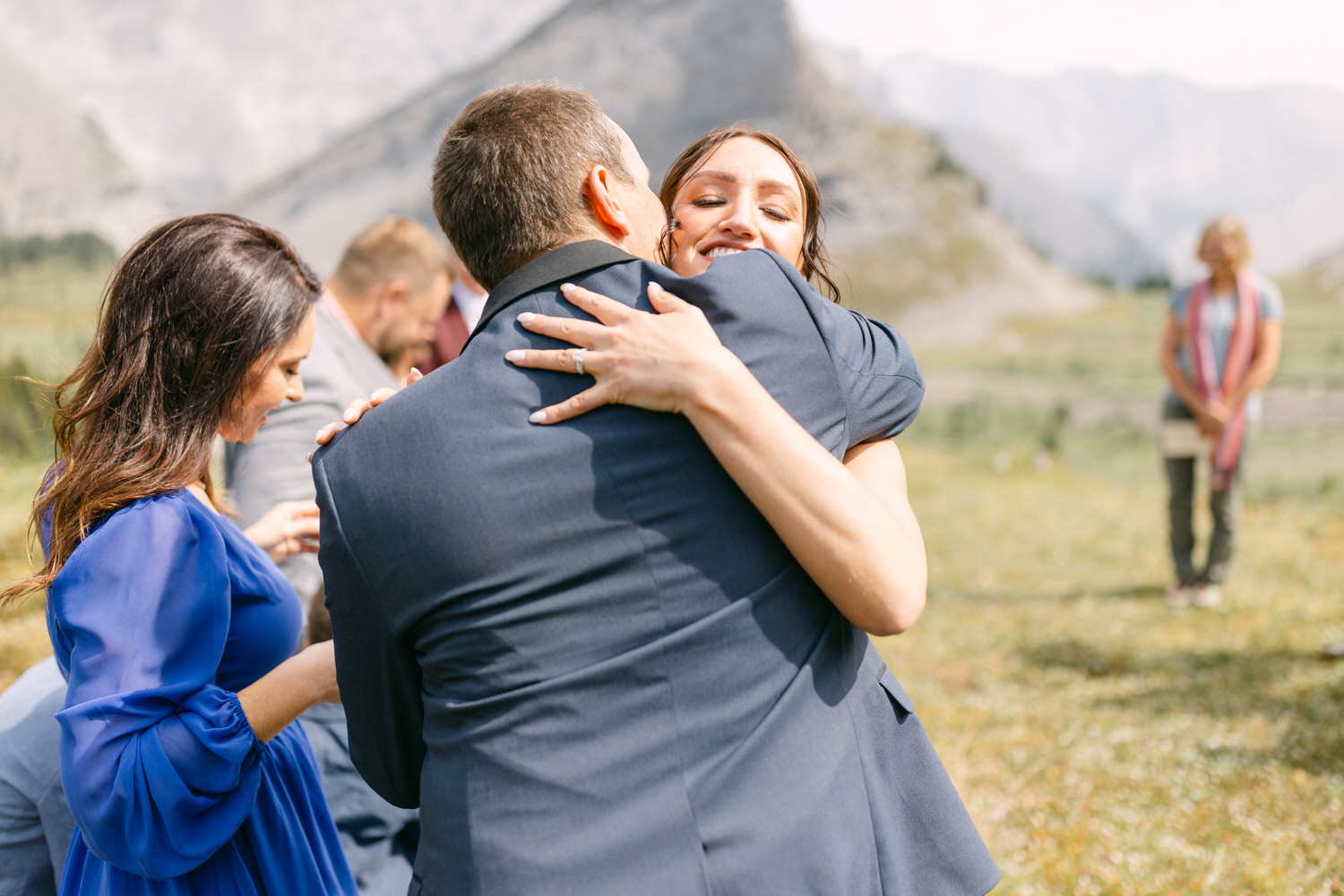 A joyful embrace between two people in formal attire, surrounded by a scenic outdoor backdrop, with others in the background engaging with their phones.