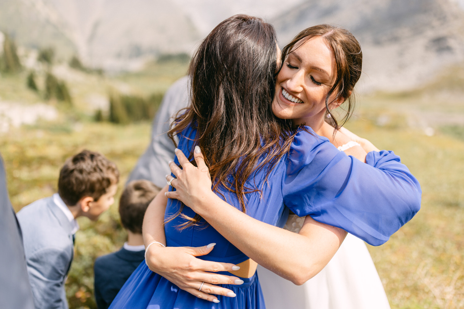 Two women share a heartfelt hug in a scenic outdoor setting, surrounded by the beauty of nature and a few children in the background.