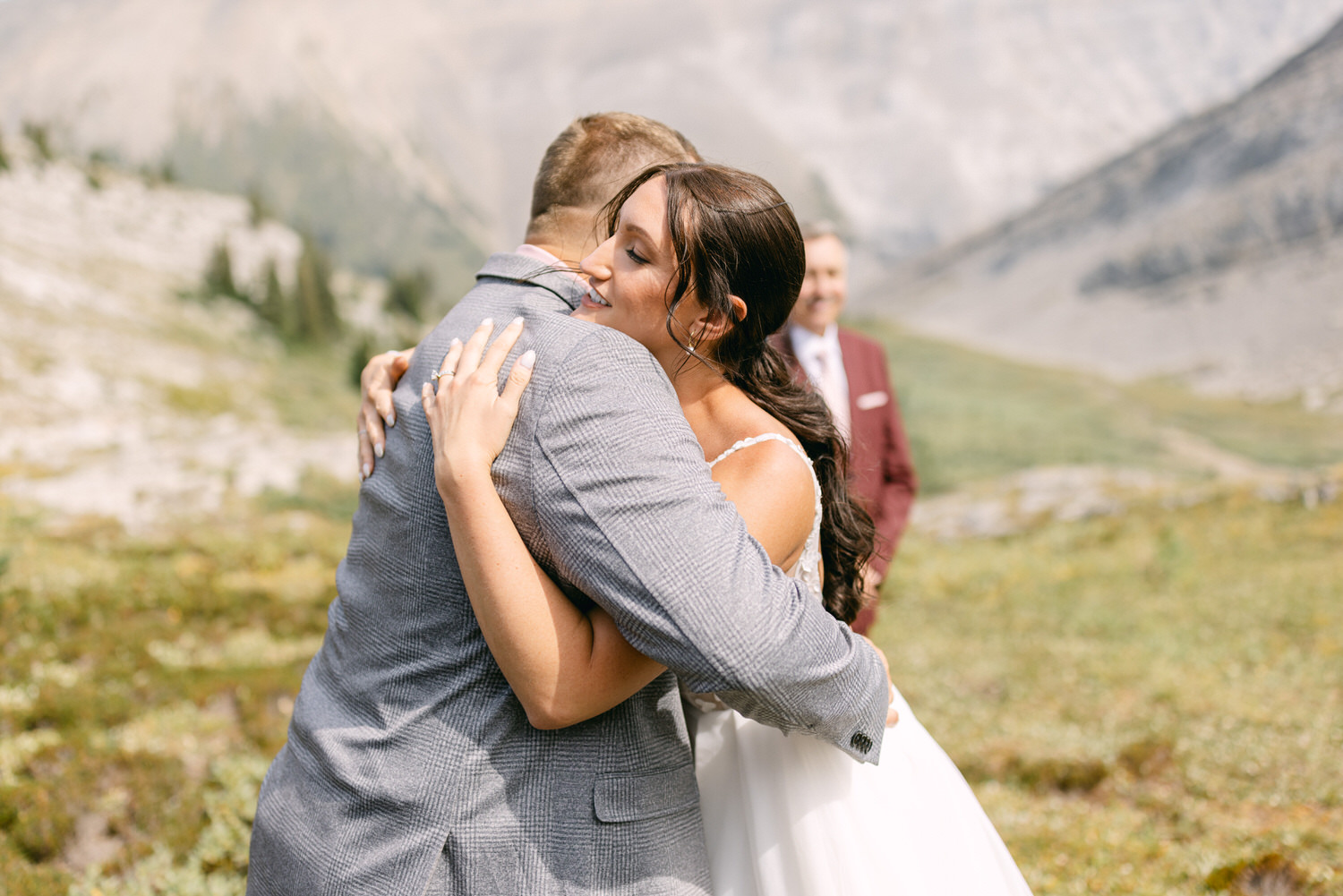 A woman in a wedding dress hugs a man in a gray suit, surrounded by a scenic mountain landscape, with another person in a burgundy suit in the background.