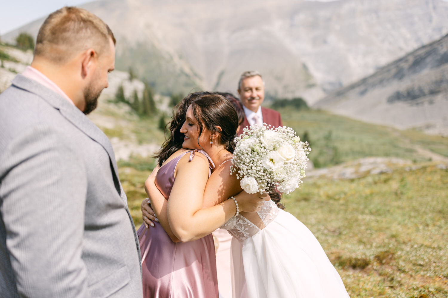 Friends sharing a joyful hug amidst a picturesque outdoor wedding setting, with mountains in the background and floral arrangements in hand.