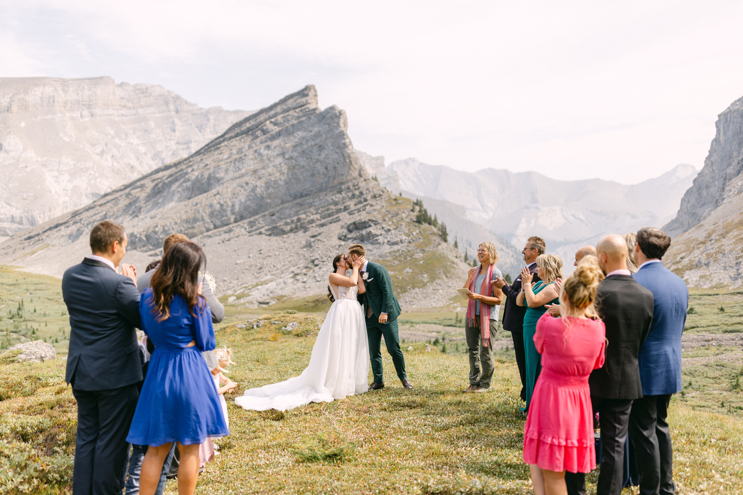 A couple shares a kiss during their outdoor wedding ceremony with a backdrop of majestic mountains and a gathering of guests.