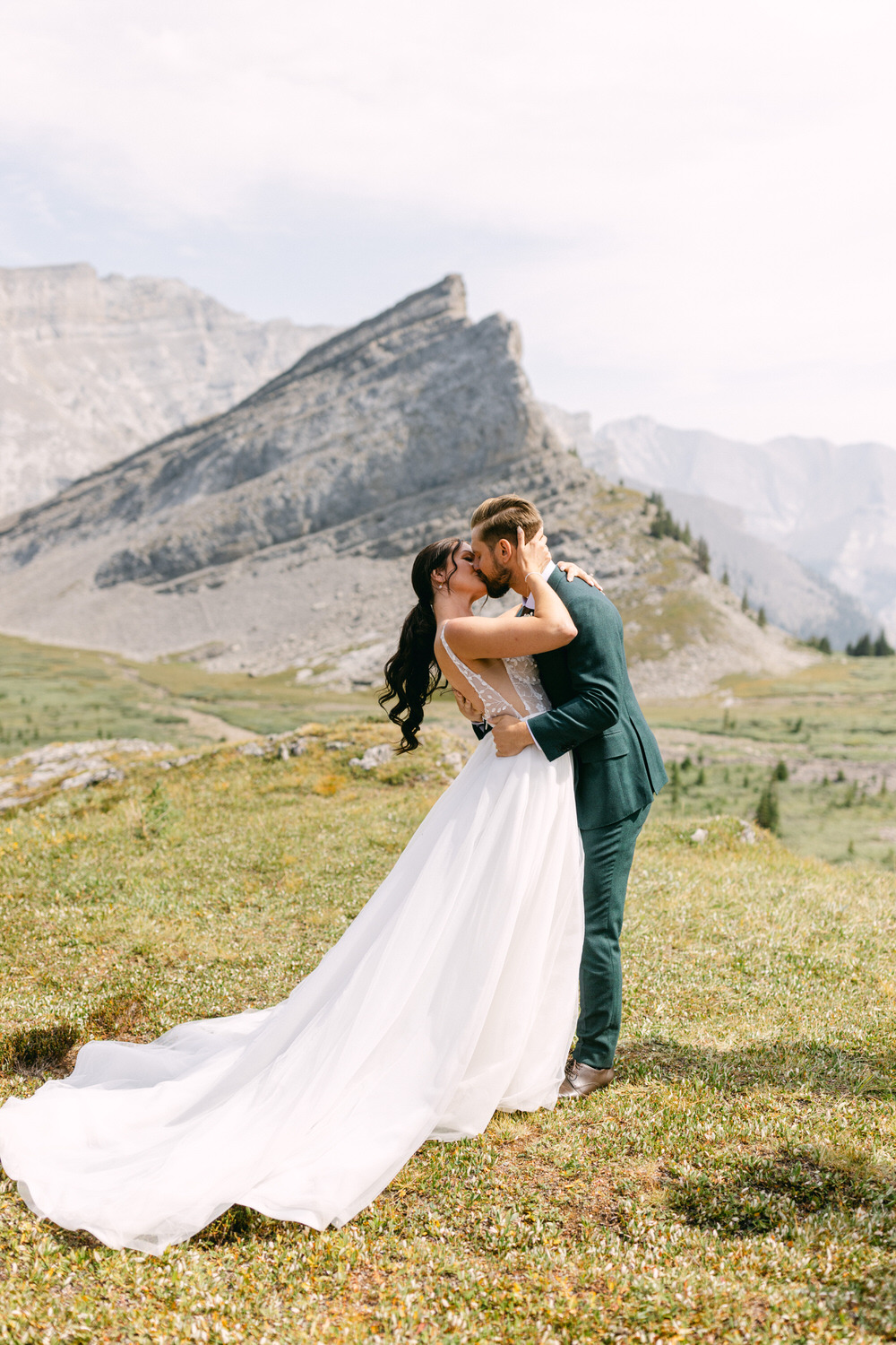 A couple embraces in a passionate kiss on a grassy mountain landscape, with breathtaking peaks in the background, celebrating their love.