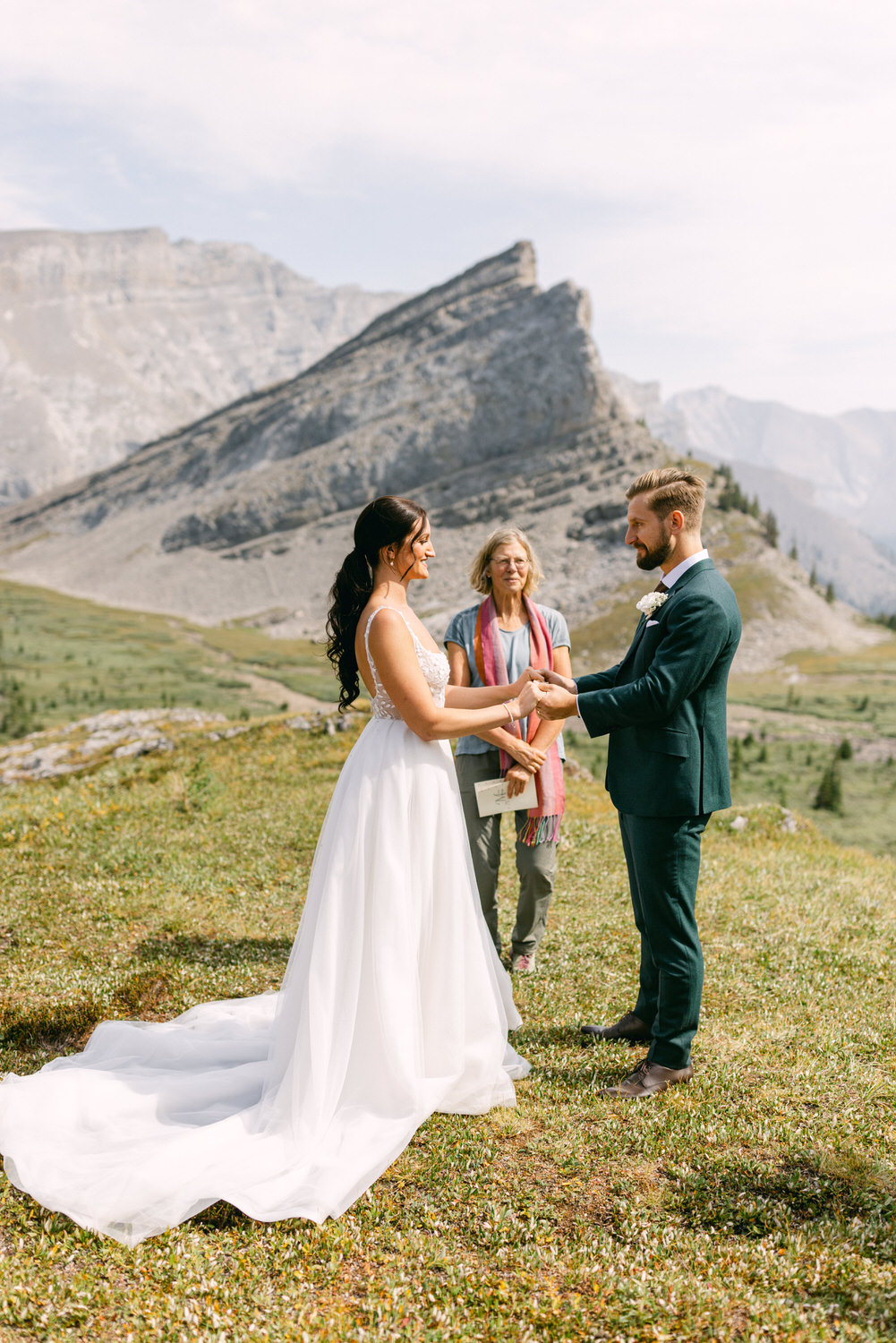 A couple exchanges vows during their outdoor wedding against a backdrop of mountains, with an officiant present.