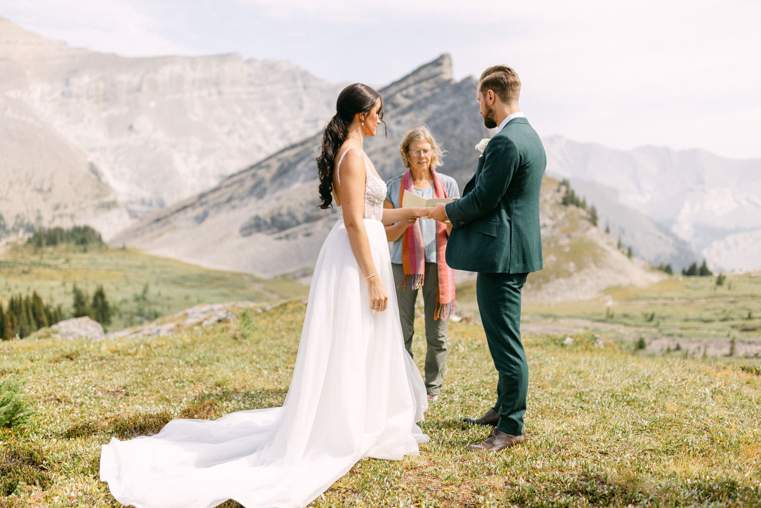 A couple exchanges vows during a picturesque outdoor wedding ceremony in a scenic mountain landscape, with a celebrant officiating.