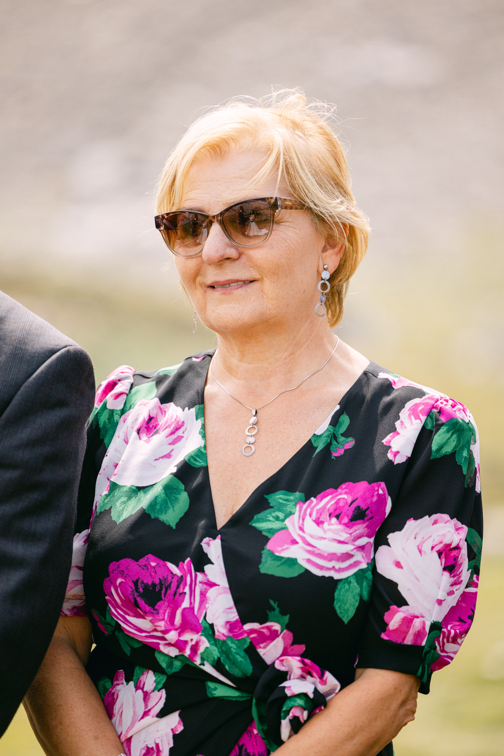 A woman with short blonde hair, wearing sunglasses and a floral-patterned dress, stands outdoors with a serene expression. She is accessorized with a silver necklace and earrings, showcasing her stylish appearance.