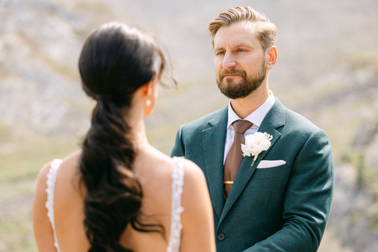 A man in a green suit with a white flower boutonniere gazes at a woman during their outdoor wedding ceremony.