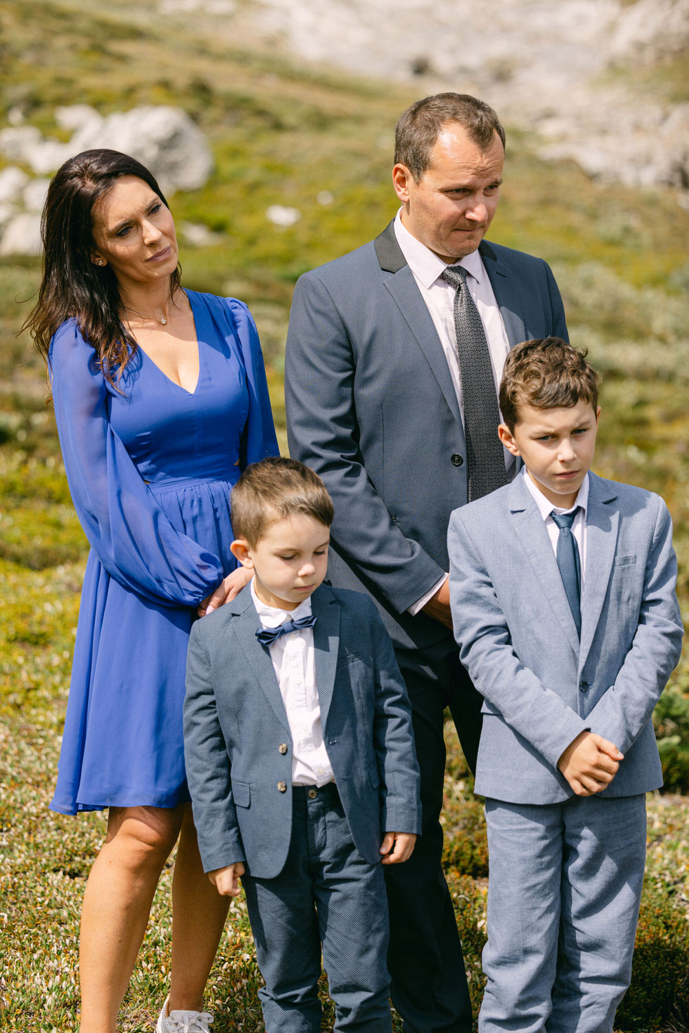 A family of four stands together in a serene outdoor setting, dressed in formal attire, with expressions of contemplation.