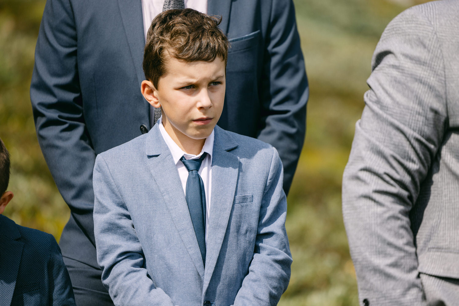 A young boy wearing a light blue suit and tie stands with a serious expression, surrounded by adults in formal attire during a sunny outdoor gathering.