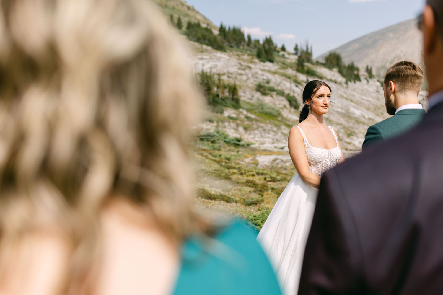 A bride in a white dress stands outdoors, surrounded by natural scenery, while looking towards her partner during their wedding ceremony. Blurry figures of guests can be seen in the foreground.