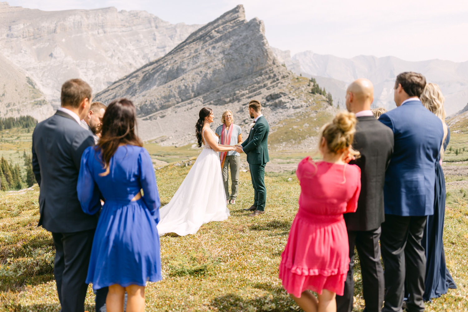 A couple exchanges vows at an outdoor wedding ceremony in a mountainous landscape, surrounded by their guests.