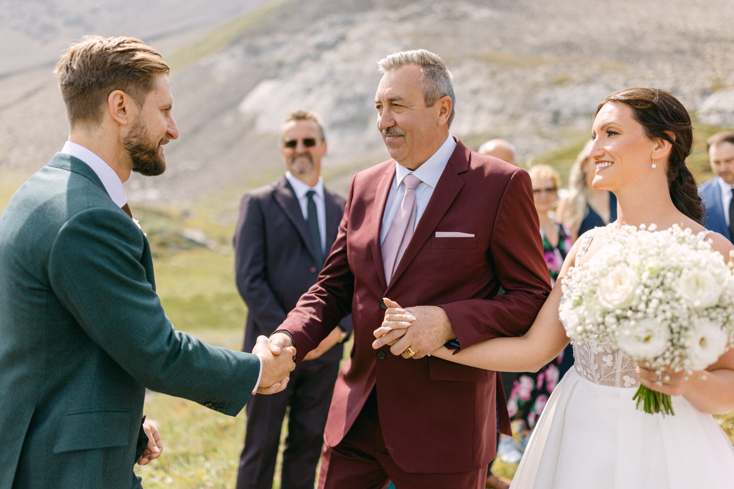 A joyful outdoor wedding scene where the groom shakes hands with the bride's father, while the bride smiles holding a bouquet of flowers.