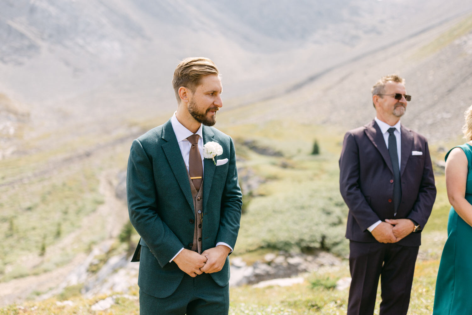 A groom in a teal suit with a white flower boutonniere stands quietly, flanked by guests, against a scenic mountain backdrop.