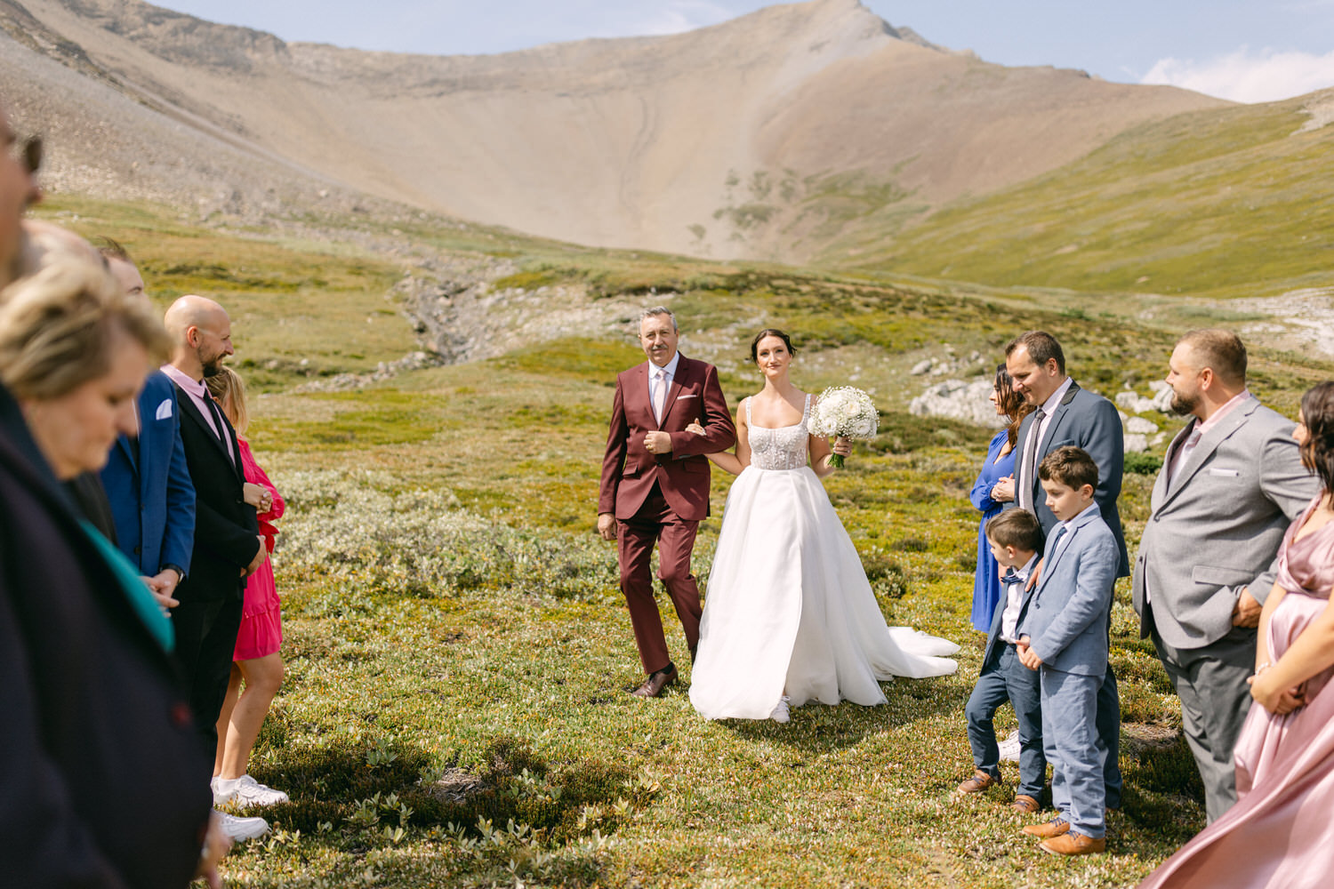 A beautiful bride in a white gown walks with her father towards a gathering of guests in a scenic mountain setting, surrounded by lush greenery and rocky terrain.