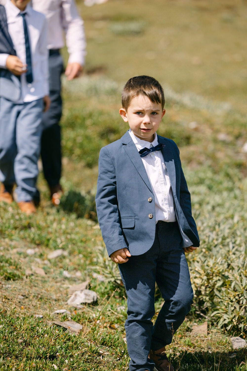 A young boy dressed in a stylish suit and bow tie stands on a grassy field, looking towards the camera while other children walk in the background.