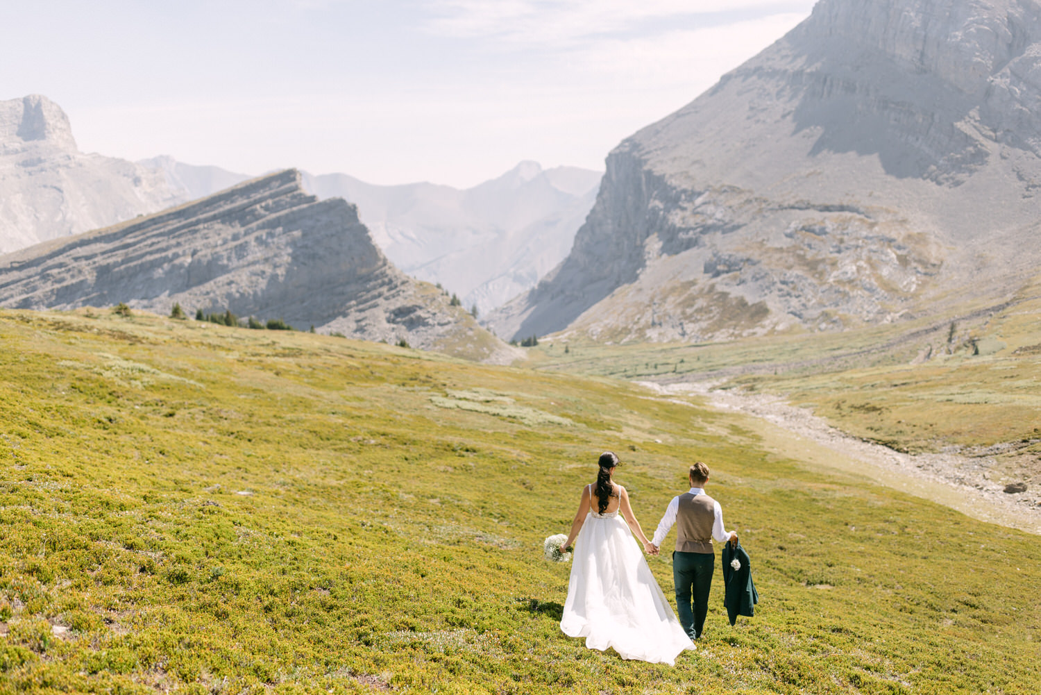 A couple holding hands, walking through a vibrant green meadow surrounded by mountains, with the bride in a flowing white dress and the groom in a vest and trousers.