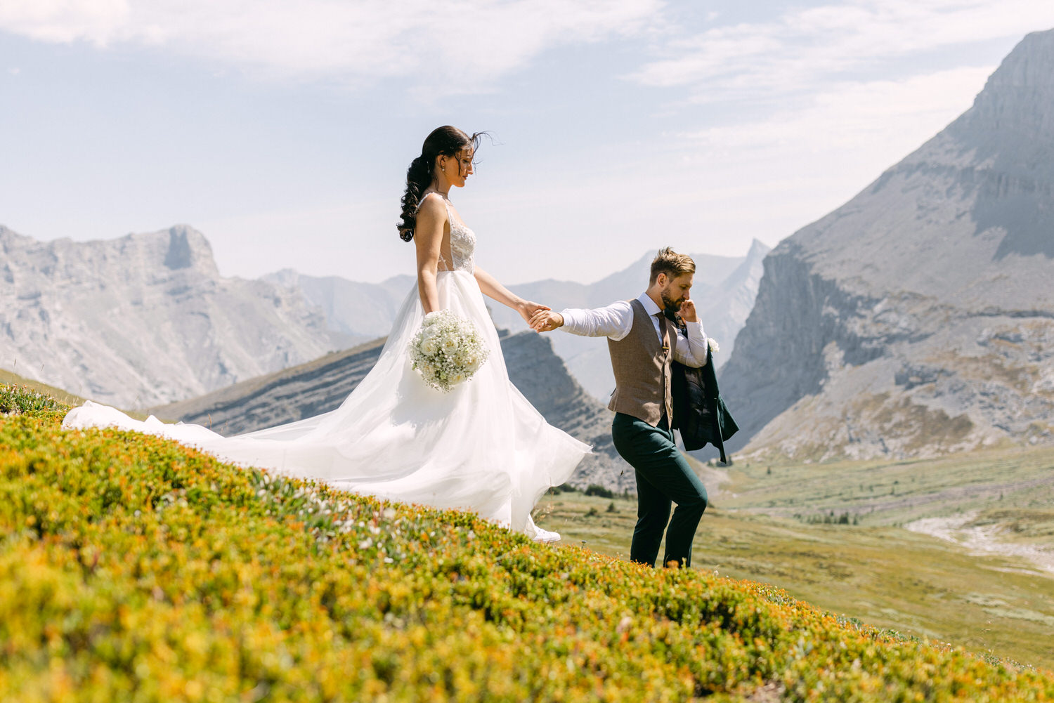 A bride and groom walk hand-in-hand through a vibrant landscape, surrounded by majestic mountains.