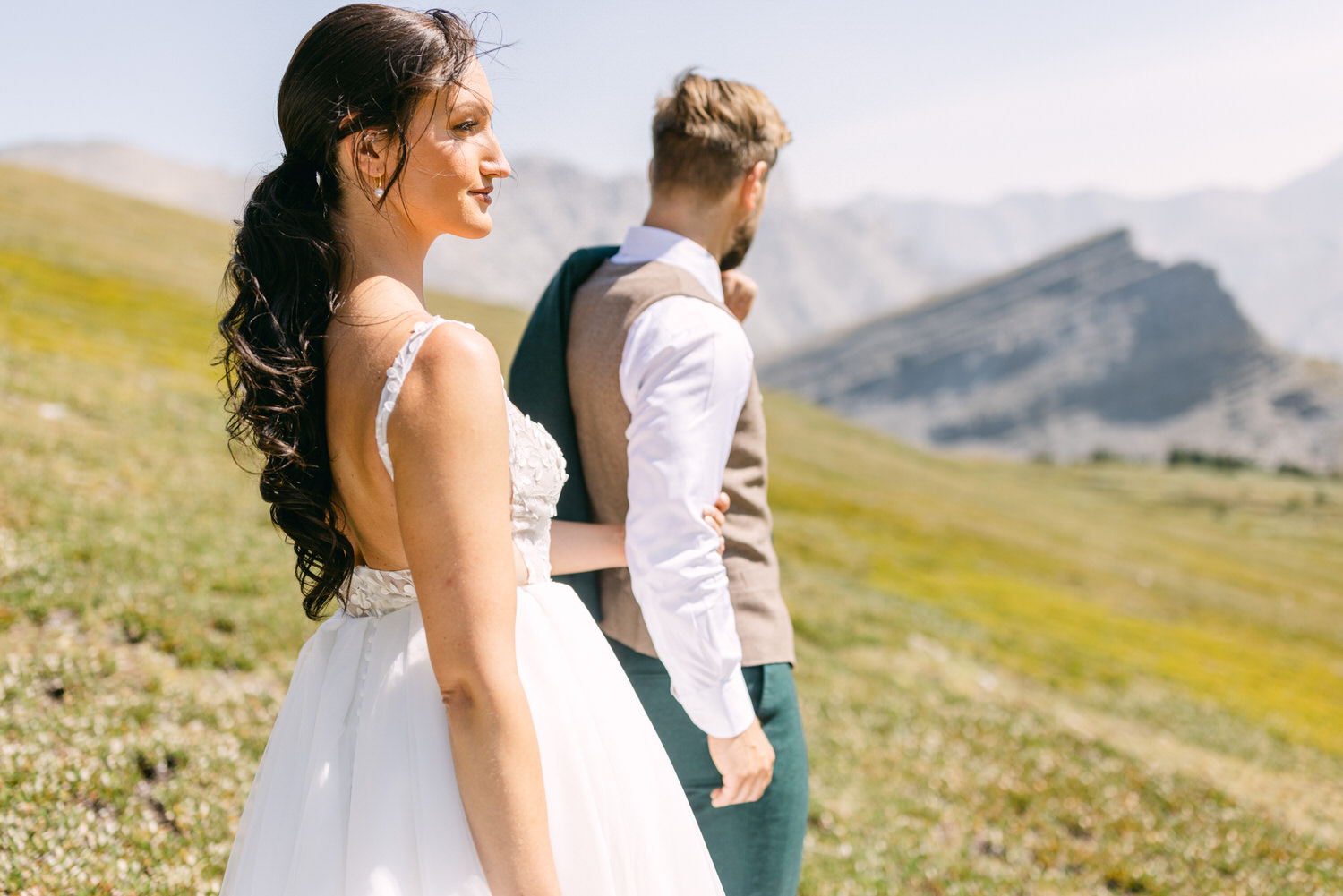 A bride with long hair in a white dress shares a tender moment with a groom in a vest as they walk through a picturesque mountainous landscape.