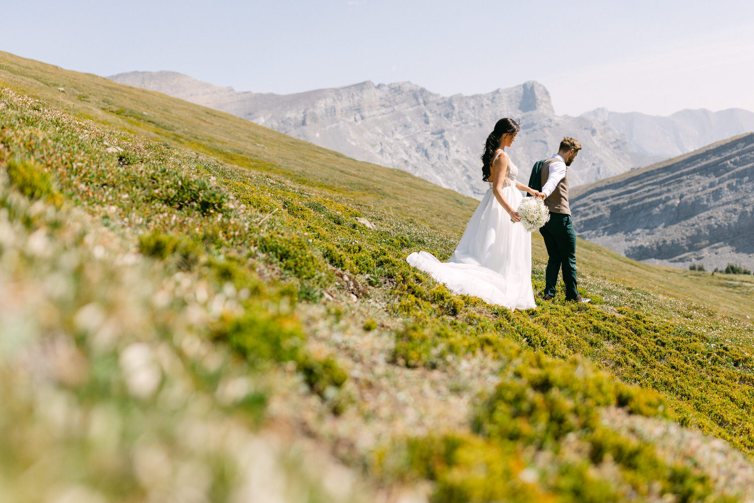 A couple in formal attire walking hand in hand through a scenic mountainous landscape, with the bride holding a bouquet and wearing a flowing white gown.