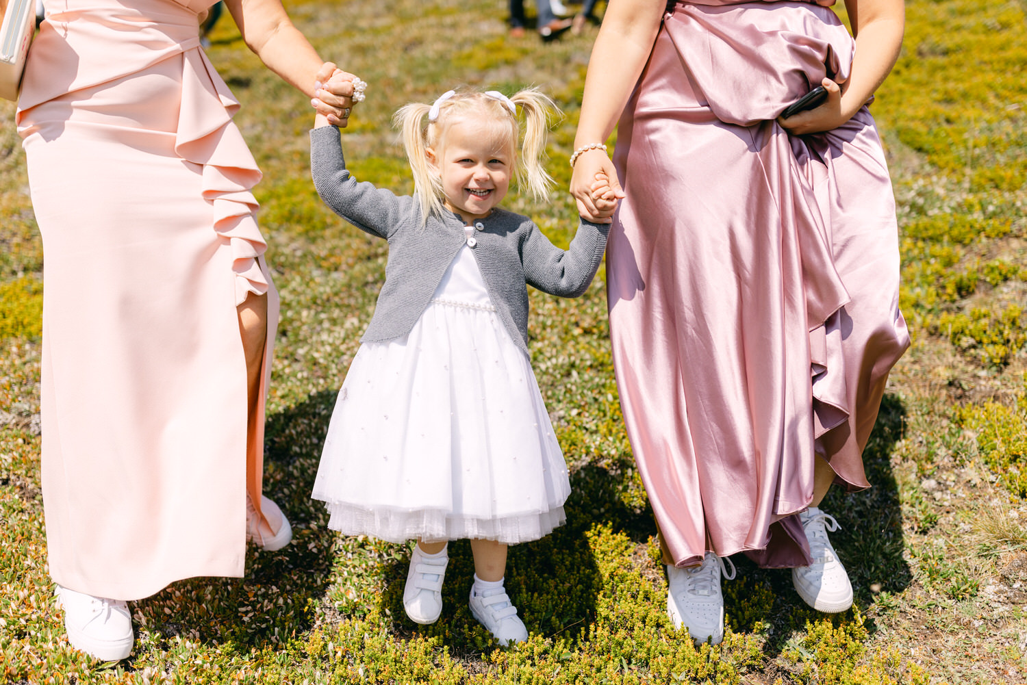 A happy little girl in a white dress and gray sweater holding hands with two adults in elegant dresses, standing on a grassy field.