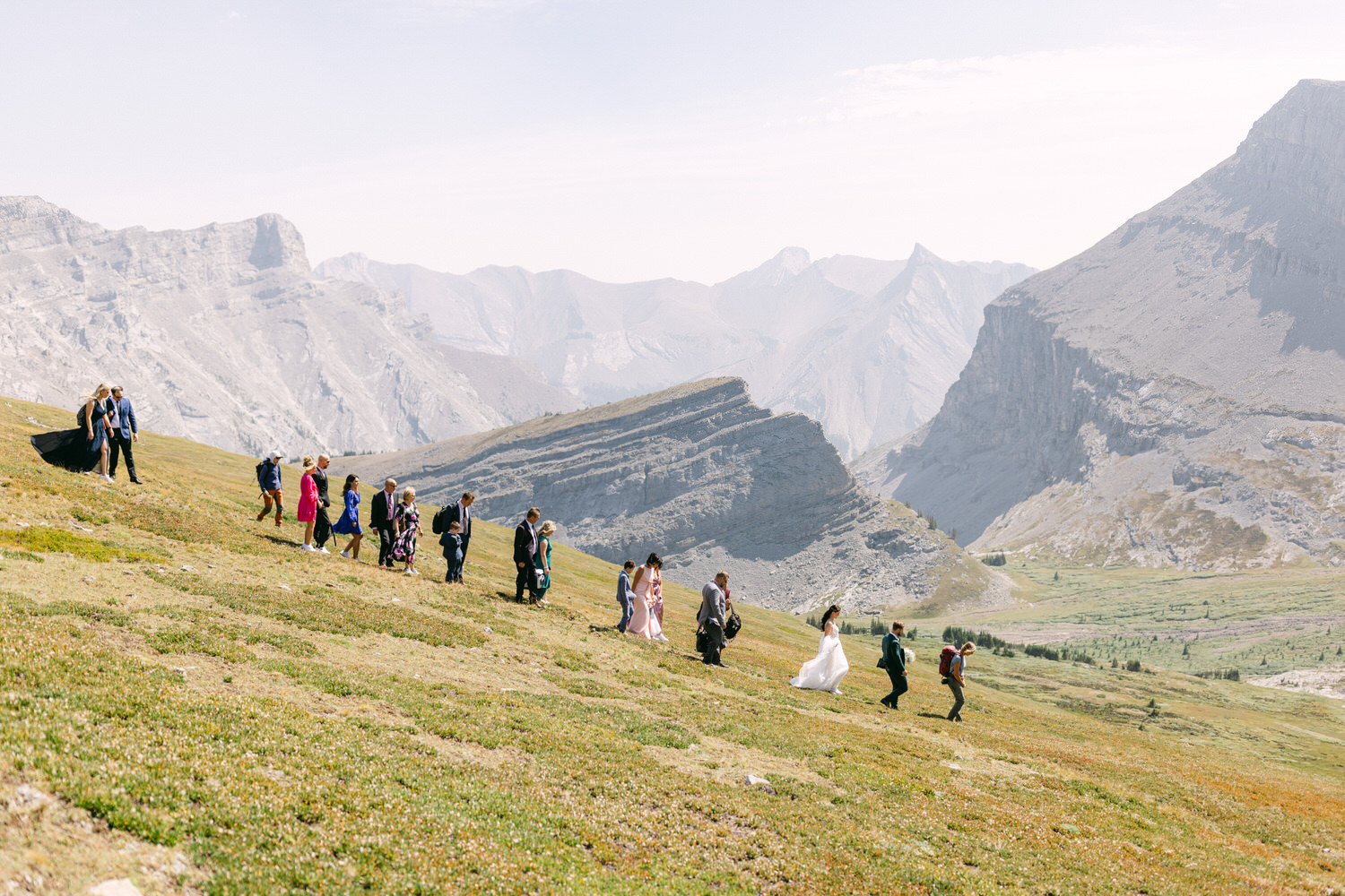 A group of wedding guests walking up a scenic mountainside, with the bride in a white dress leading the way, surrounded by beautiful valleys and peaks in the background.