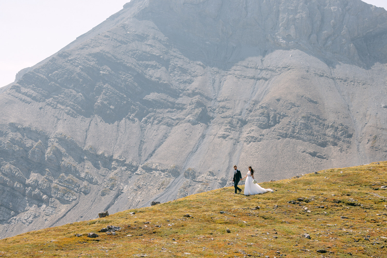 A couple walks hand in hand on a grassy hilltop, surrounded by majestic mountains in the background.