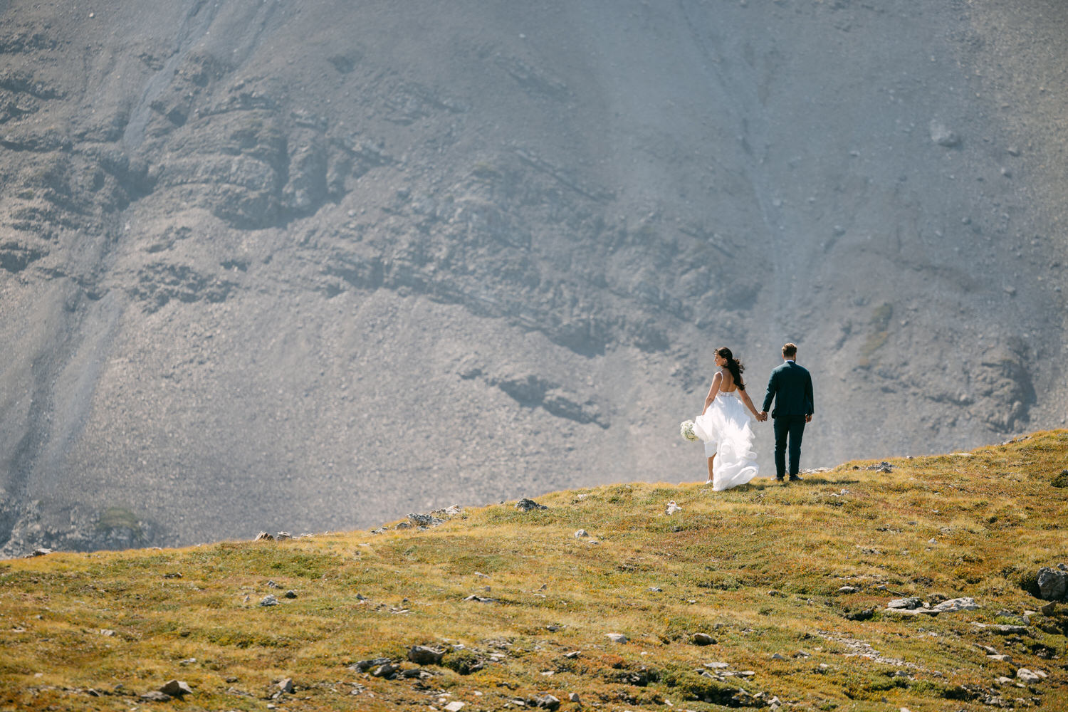 A couple strolls hand-in-hand on a grassy hilltop with a beautiful mountainous backdrop on their wedding day.