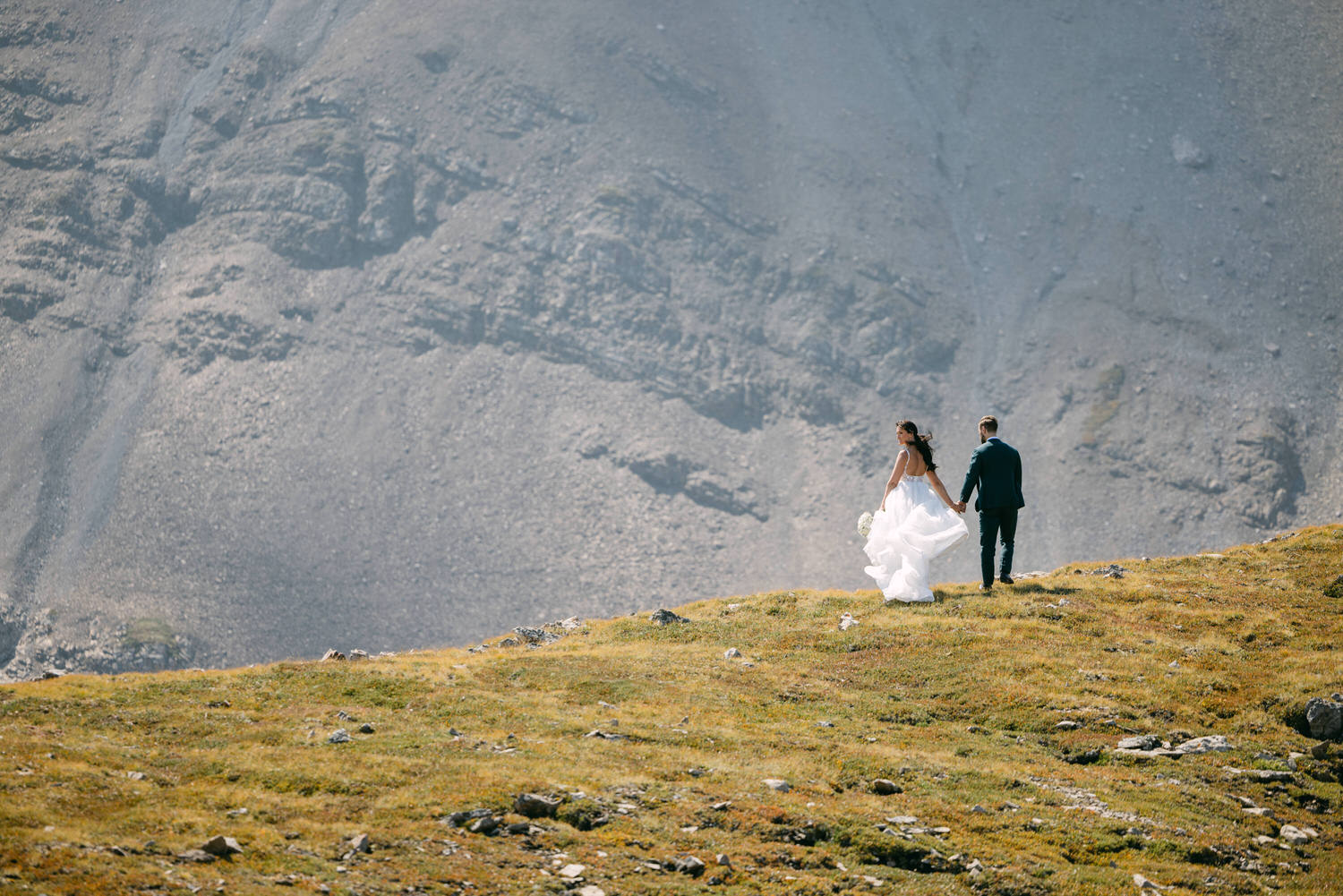 A couple in formal attire walking hand in hand on a grassy hillside with rocky mountains in the background.